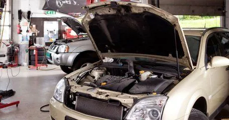 A car with its bonnet up inside the workshop, a second car behind it and the roller door open to the yard.