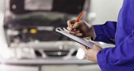 A technician in blue overalls writing on a clipboard beside a car with its bonnet up.